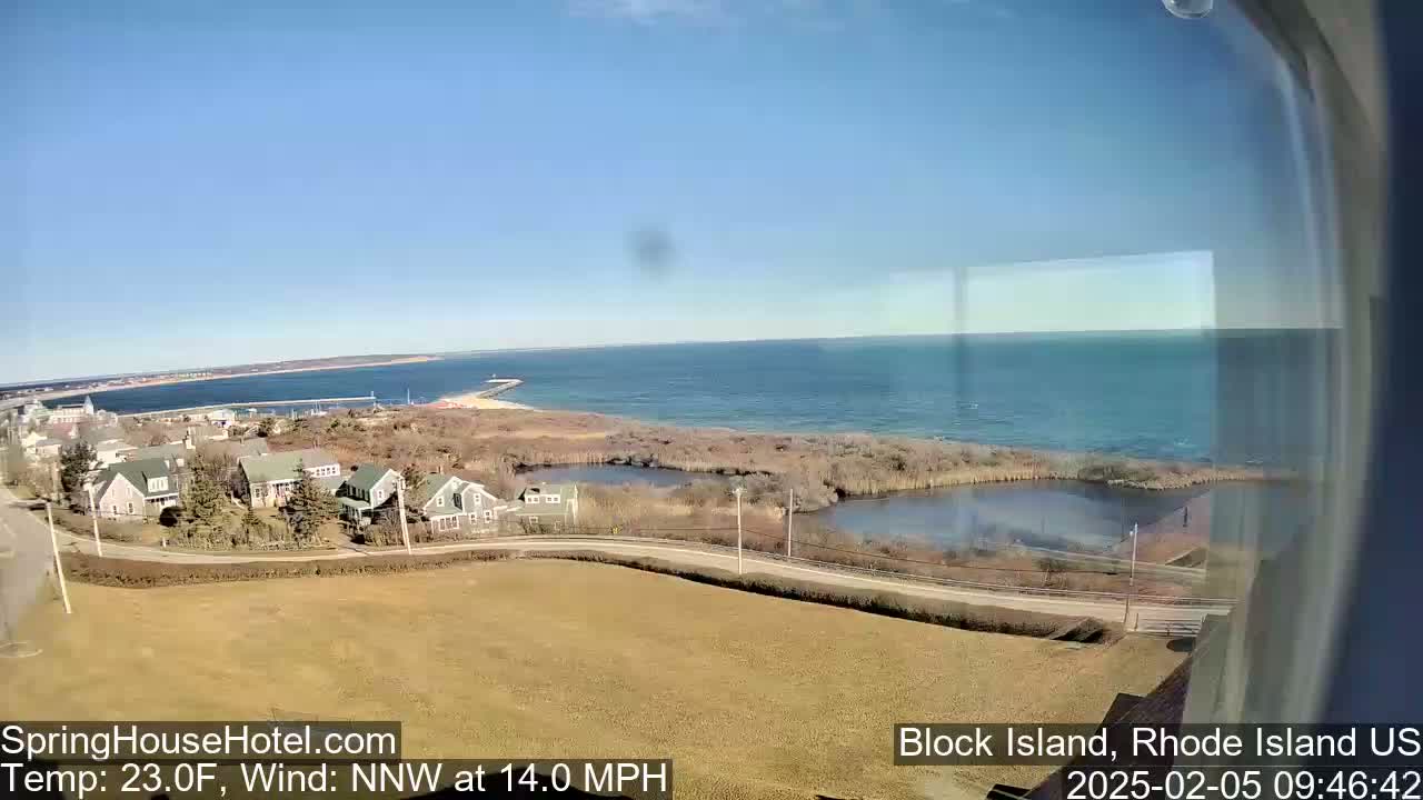 A coastal view on a clear, sunny day shows a residential area near a calm ocean with a small body of water between the houses and the shore.