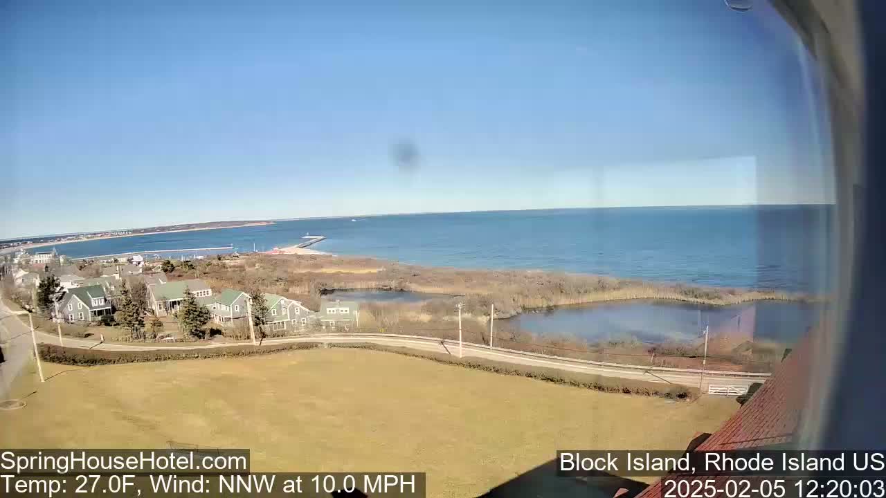 A clear, sunny day shows a coastal view of houses, a road, and a calm body of water with a distant horizon.