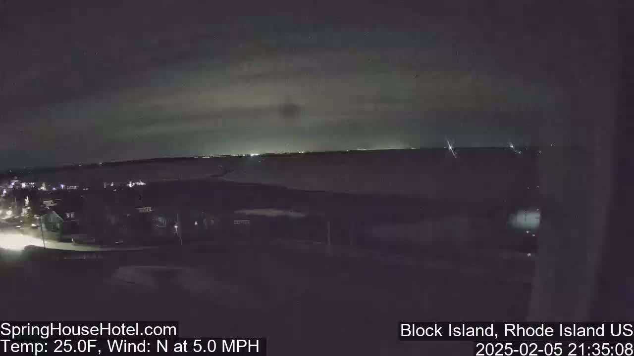 A nighttime, overhead view of a town near a body of water under a partly cloudy sky with a north wind blowing at 5.0 mph.