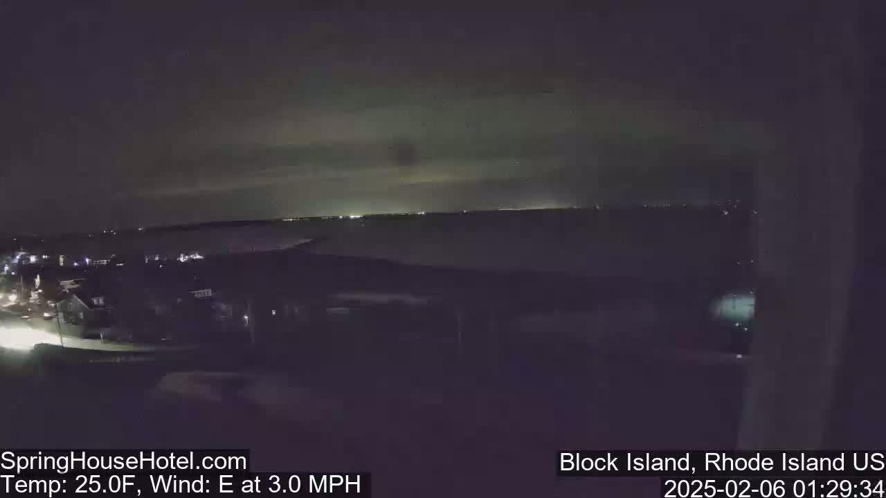 A nighttime, long shot of a dark body of water with faintly visible lights on the opposite shore, under a slightly cloudy sky, with a light east wind.
