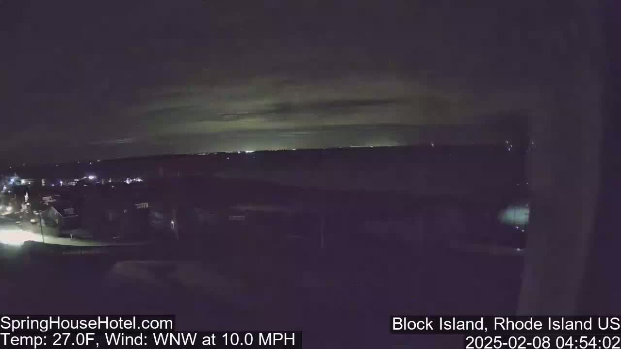 A nighttime, long shot of a town with faintly visible lights under a mostly cloudy sky, with a 27°F temperature and a 10 mph wind from the west-northwest.