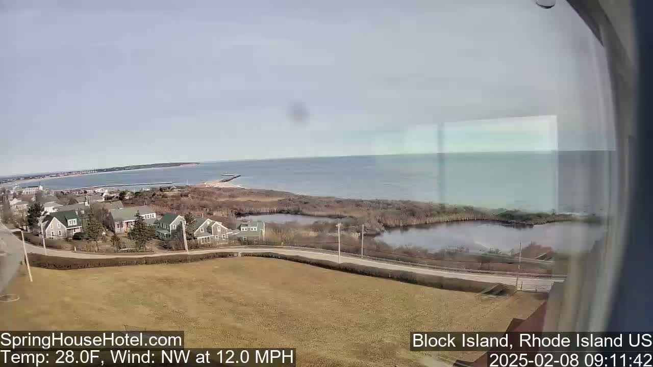 A coastal town with houses and a calm body of water is seen from a high vantage point on a partly sunny day with a northwest wind.