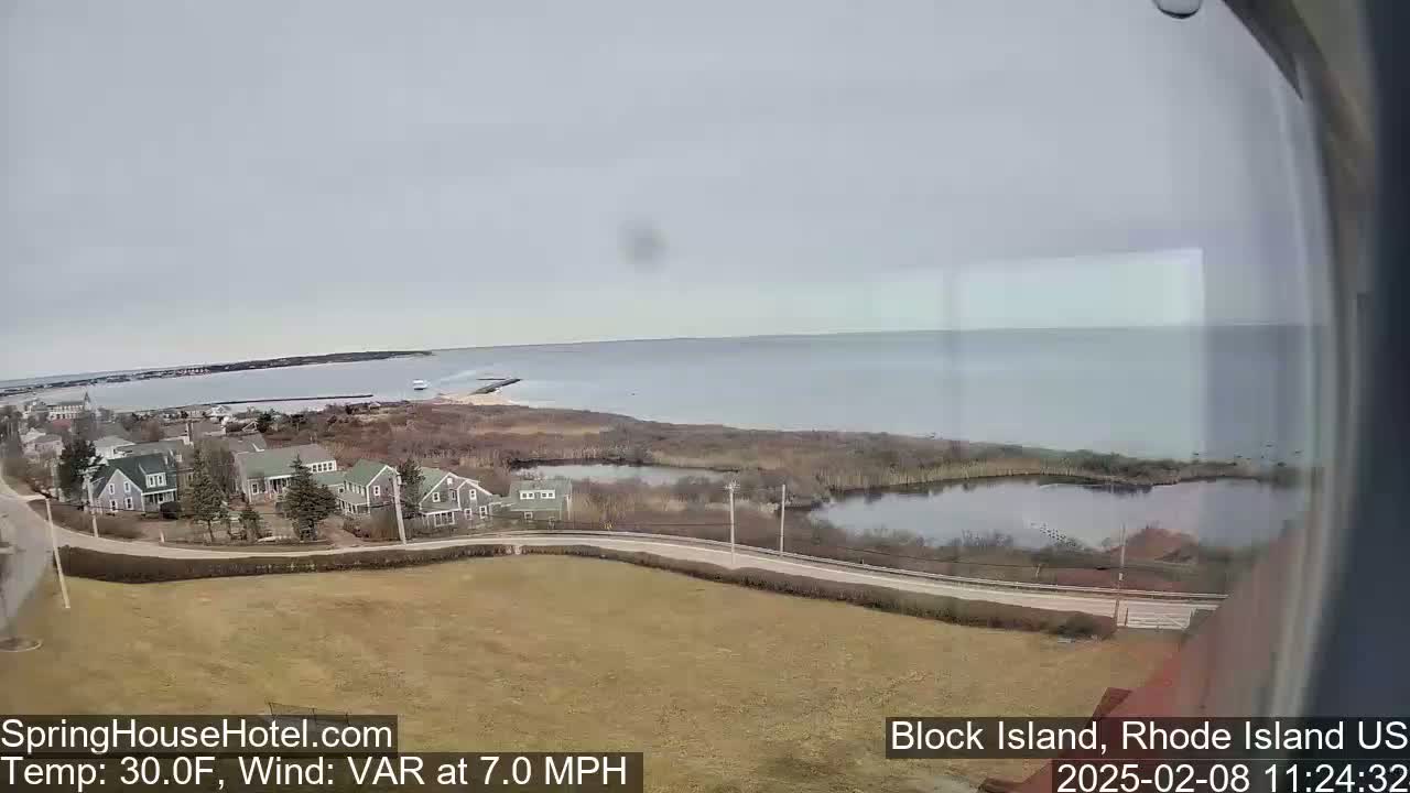 A coastal view shows a calm body of water, a small town with houses, and a marshy area with a pond, under overcast skies.