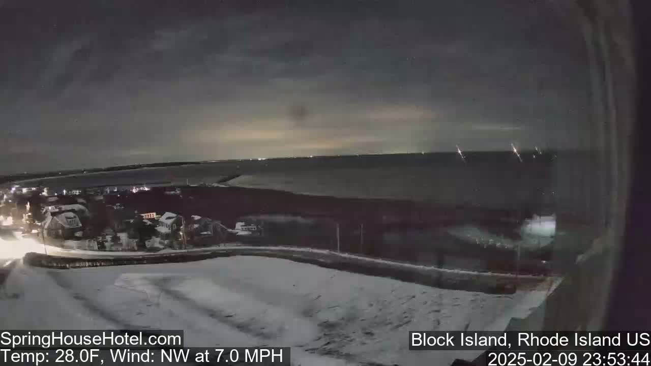 A nighttime, snow-covered landscape overlooking a dark body of water under a mostly cloudy sky with a northwest wind.