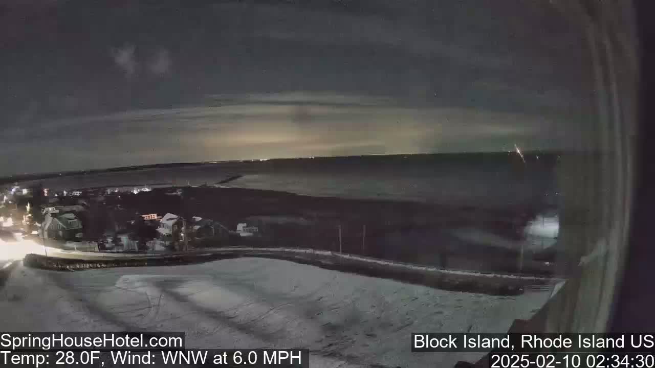 A nighttime view of a snow-covered coastal town overlooking a dark ocean under a cloudy sky with a 6 mph wind.