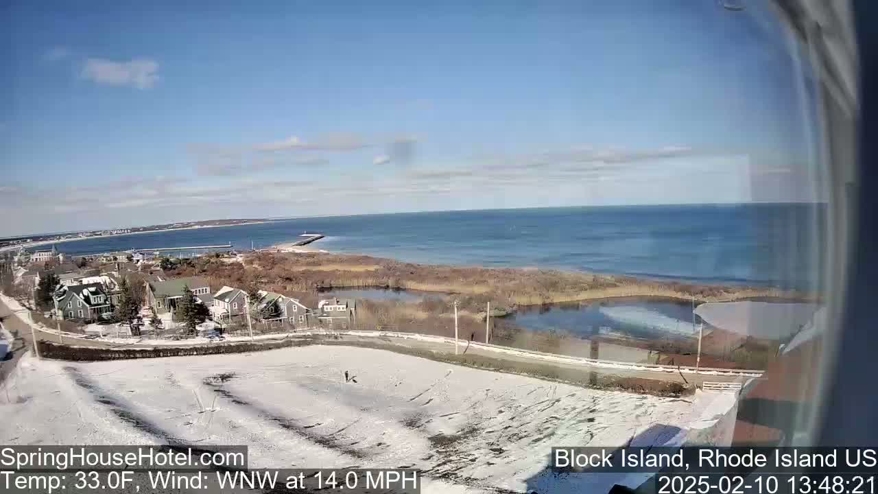 A partly sunny, cold, and windy day shows a snow-covered coastal area with houses and a body of water, partially frozen, extending to a calm ocean.