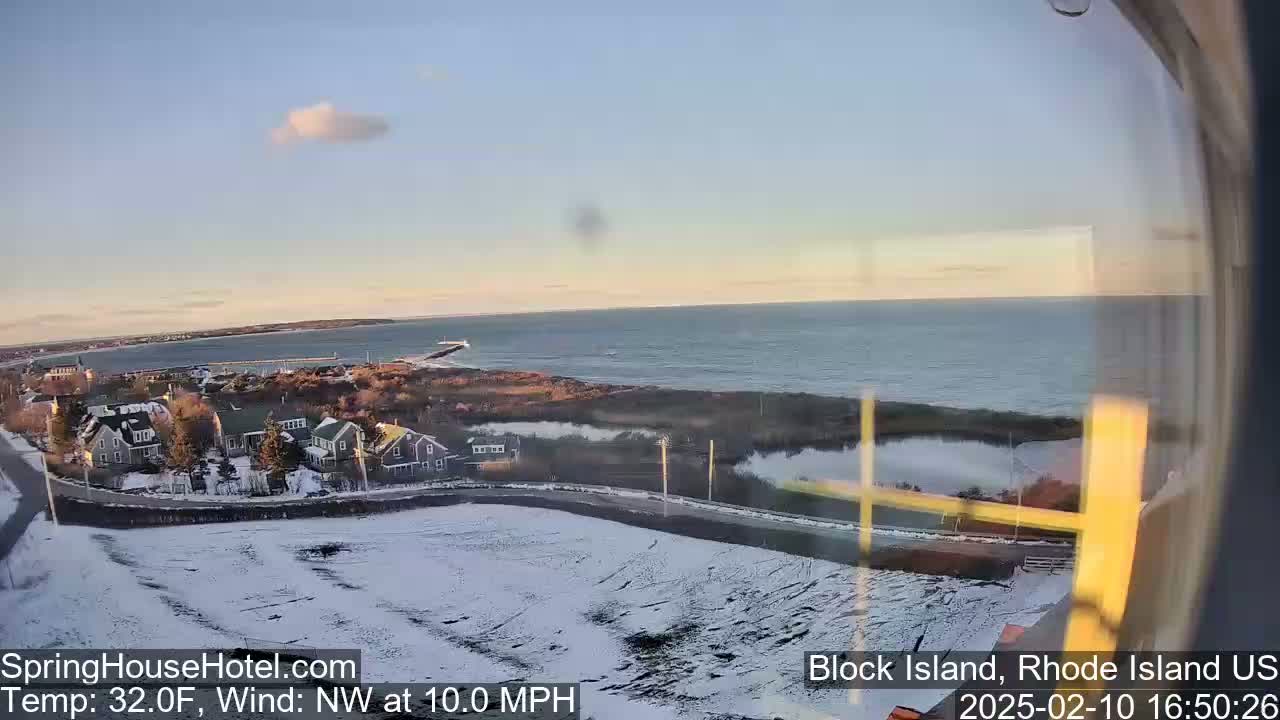 A snow-covered coastal town overlooks a calm ocean under a partly cloudy sky.