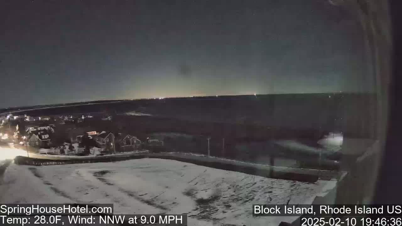 A nighttime, snowy landscape shows a residential area with a road and a body of water in the distance under a dark sky.