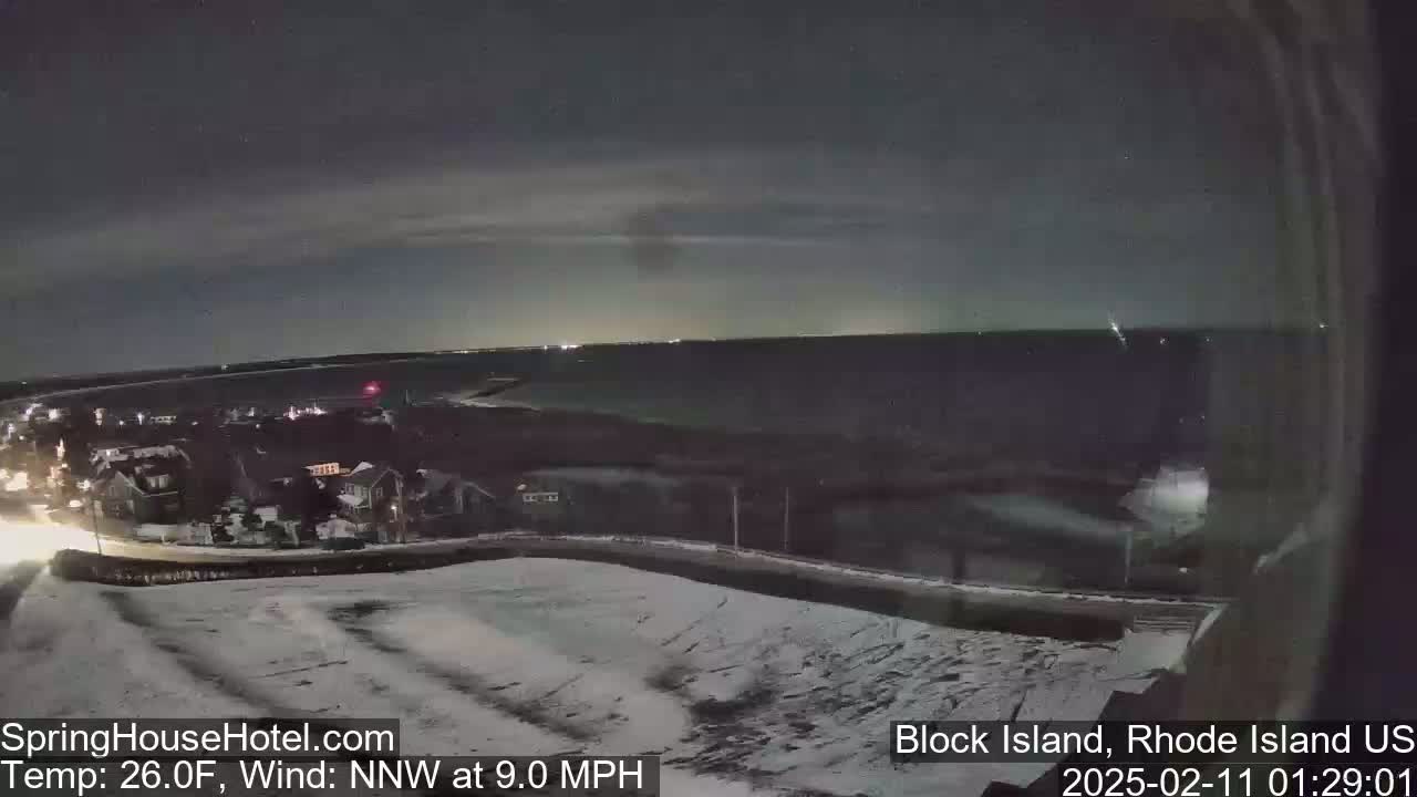 A nighttime, snow-covered landscape shows a dark body of water, residential buildings with lights, and a road under a cloudy sky with a north-northwest wind at 9.0 mph and a temperature of 26 degrees Fahrenheit.