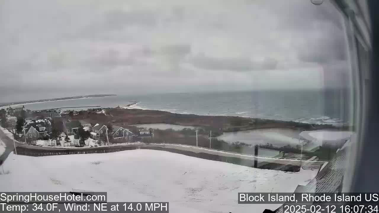 A snow-covered coastal landscape with houses and a body of water under an overcast sky, with a northeast wind blowing at 14 mph.