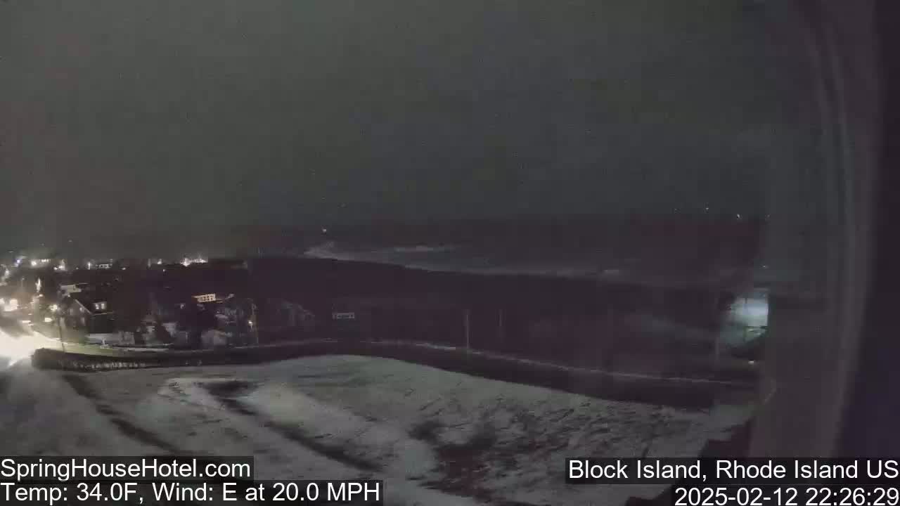 A nighttime, snowy scene shows a dark ocean with waves under a dark sky, and houses in the distance with a road in the foreground.