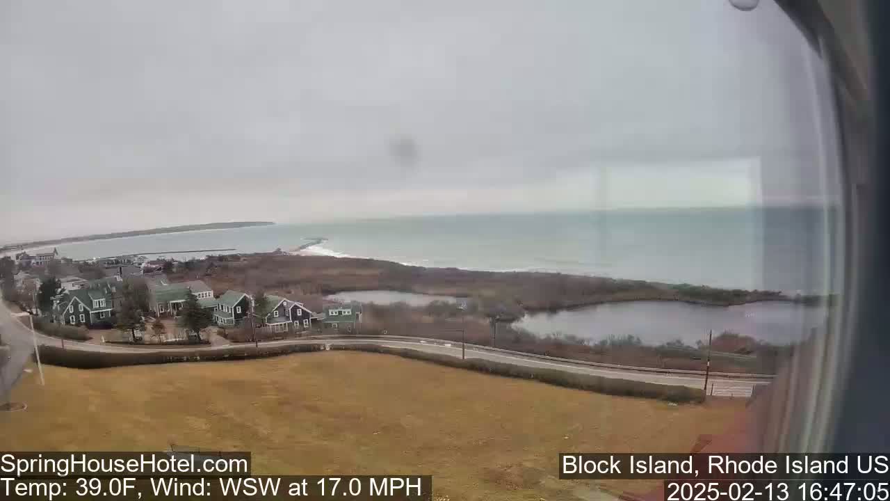 A coastal view on an overcast day shows a quiet residential area near the ocean with a calm sea, a small pond, and a road curving through the landscape.