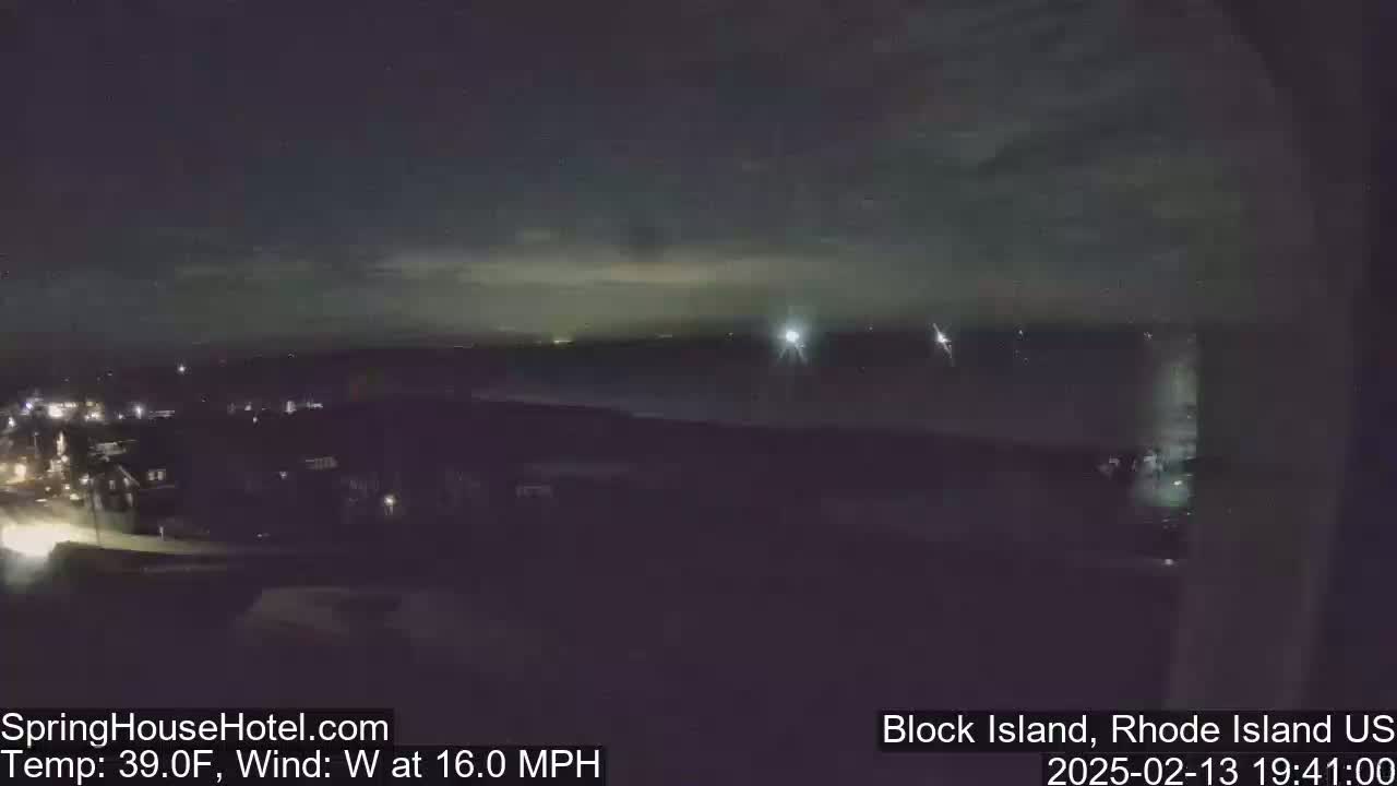 A nighttime view of a town and body of water under a dark sky with a few faint lights visible in the distance, and a 16 mph westerly wind.
