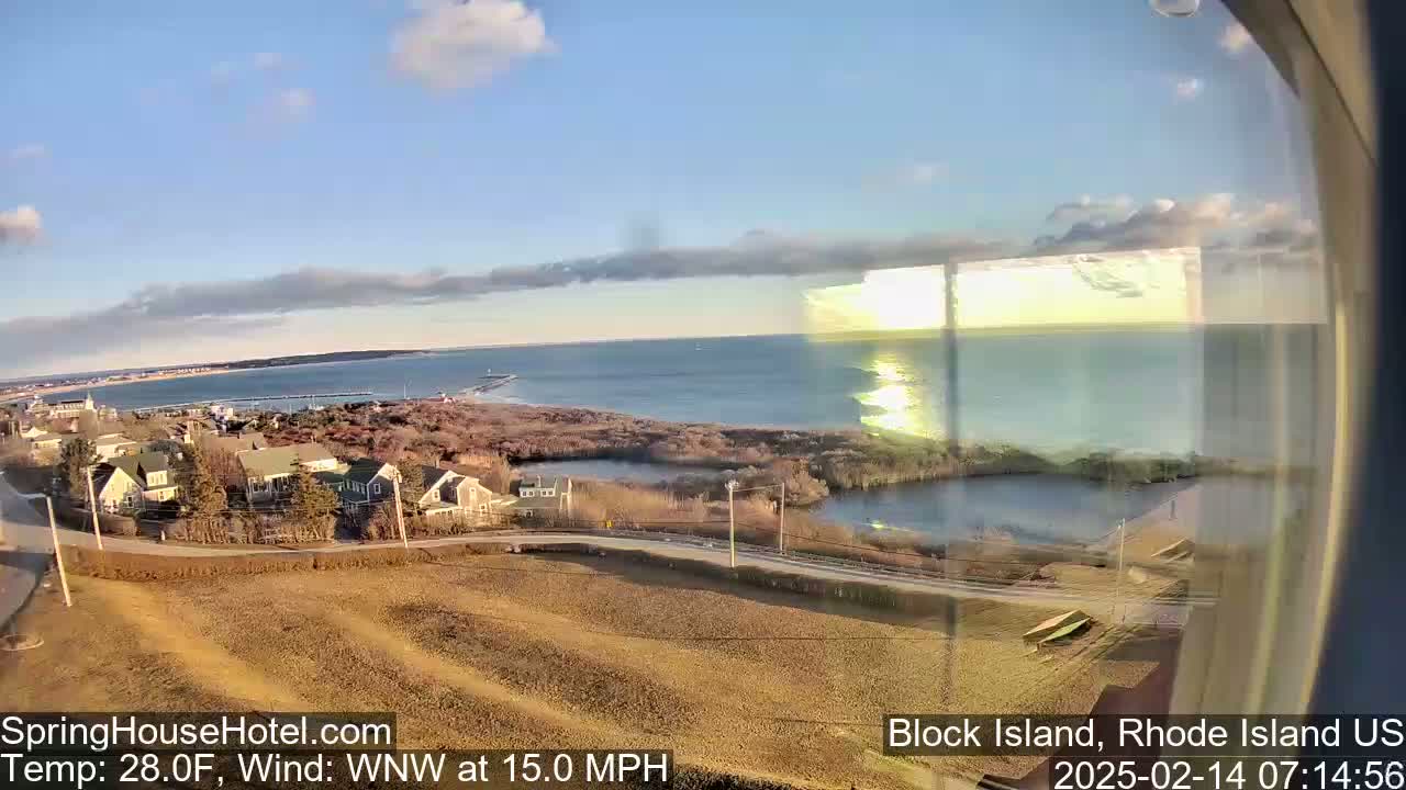 A coastal town with houses and a road overlooks a calm ocean under a partly cloudy sky, with a brisk wind.