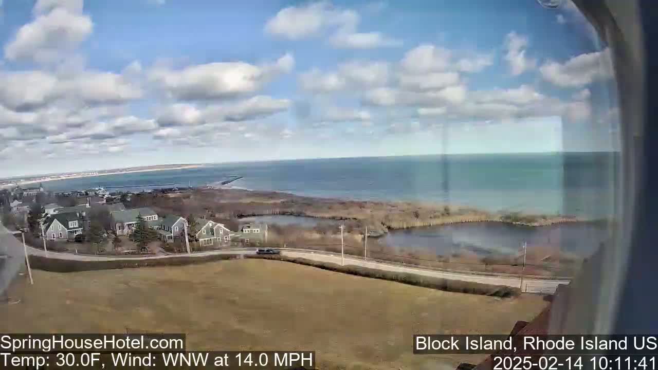 A partly cloudy day reveals a coastal scene of houses, a road, marshland, and a calm ocean under a blue sky.