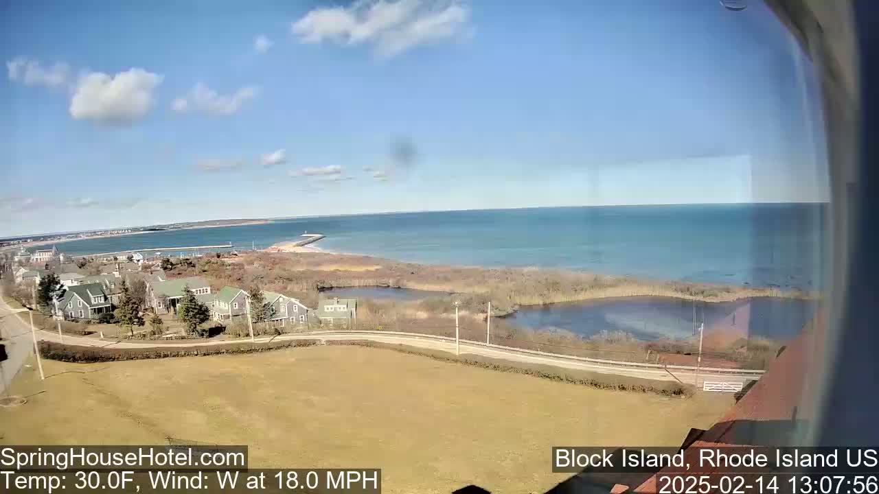 A coastal view on a partly sunny day shows a calm ocean, a sandy shoreline, houses, marshland, and a road curving along the water's edge.