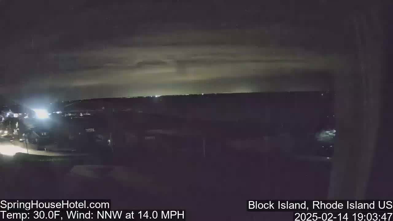 A nighttime view of a town with a body of water in the distance, under a cloudy sky with a 30°F temperature and a 14 mph northwest wind.