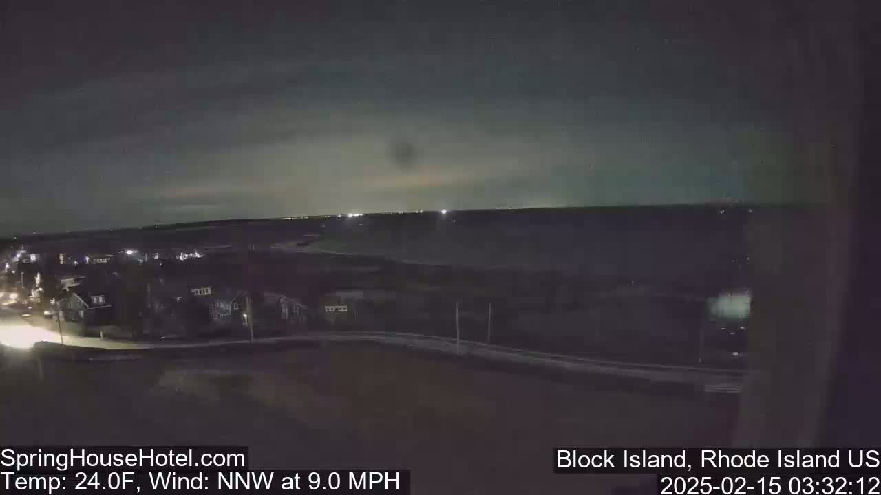 A nighttime long shot of a residential area near a body of water under a mostly clear sky with a northwest wind at 9 mph and a temperature of 24°F.