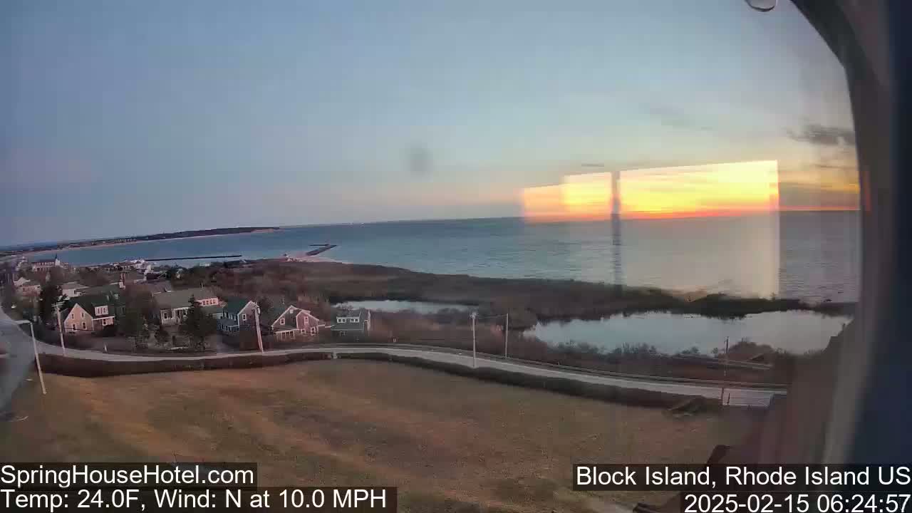 A coastal town at sunset is seen from an elevated vantage point, with a calm body of water and a north wind of 10 mph.