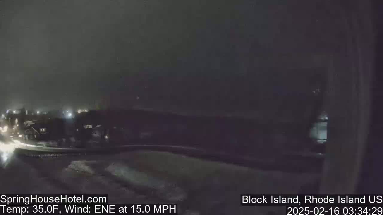A nighttime view of a snow-covered landscape with dimly lit buildings and a road, under a dark sky with a 15 mph wind from the east-northeast.