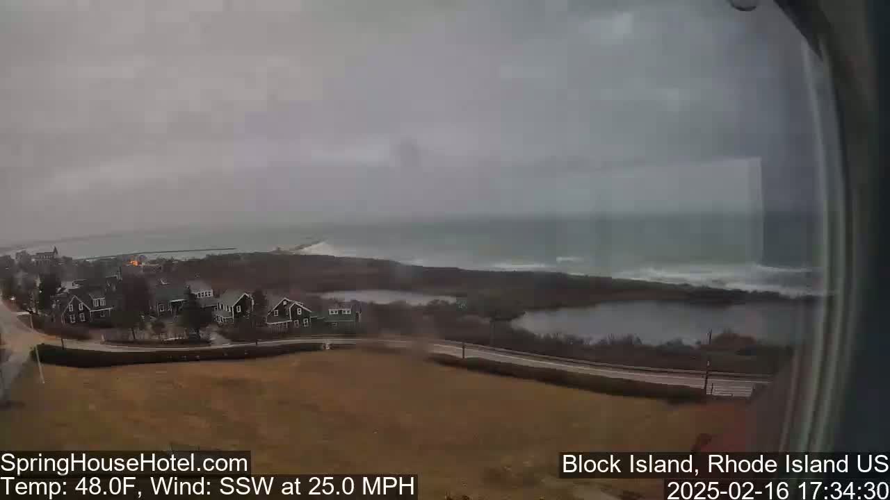 A coastal scene shows a gray, overcast day with a strong south-southwest wind; houses sit near a road overlooking a body of water with choppy waves.