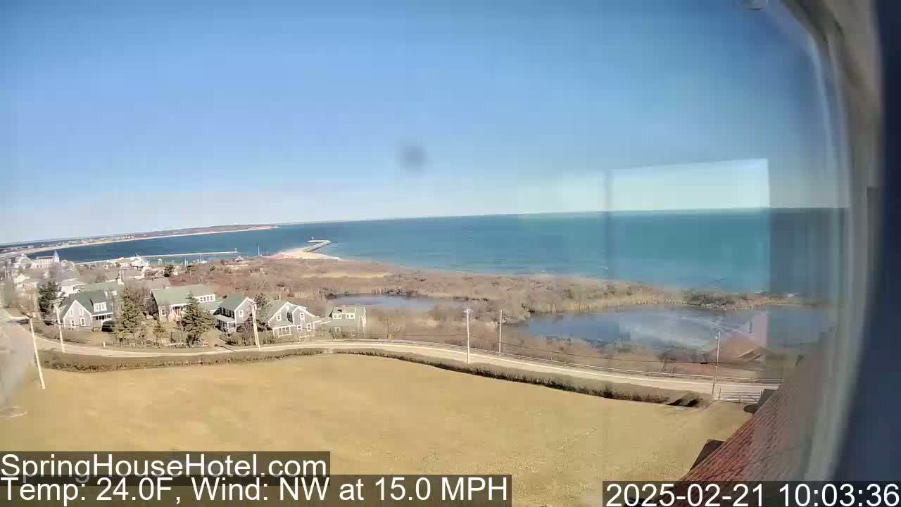 A high-angle view shows a coastal town on a sunny day with a northwest wind of 15 mph and a temperature of 24°F, overlooking a calm body of water.