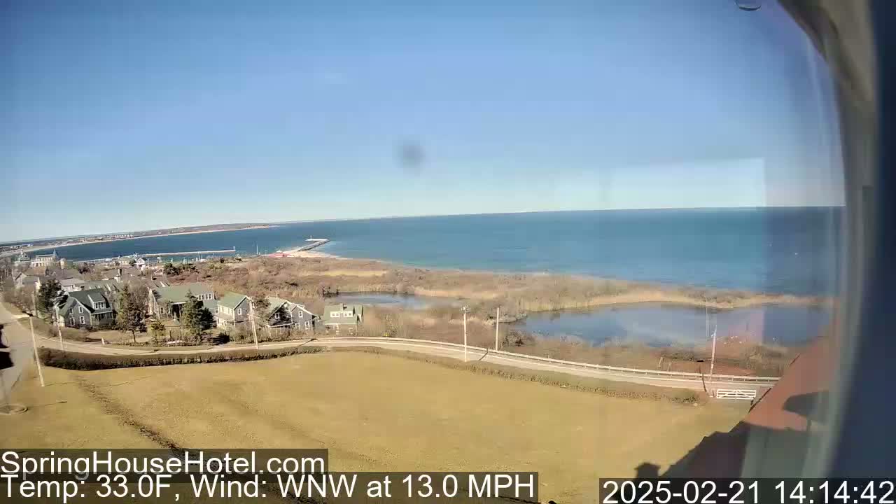 A coastal scene on a clear, sunny day with a 33°F temperature and a 13 mph wind shows a tranquil body of water, houses nestled near marshland, and a distant shoreline under a clear sky.