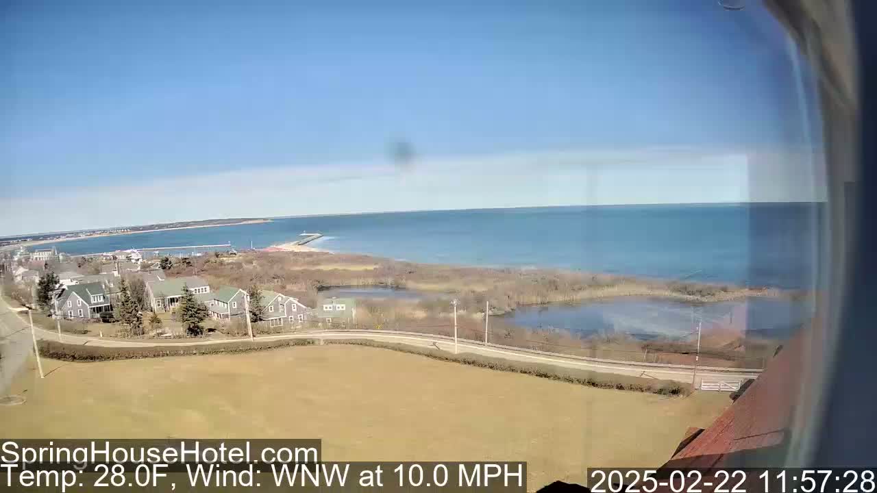 A coastal scene on a clear, sunny day with a 10 mph wind shows a body of water, a sandy beach, several houses, marshland, and a road.
