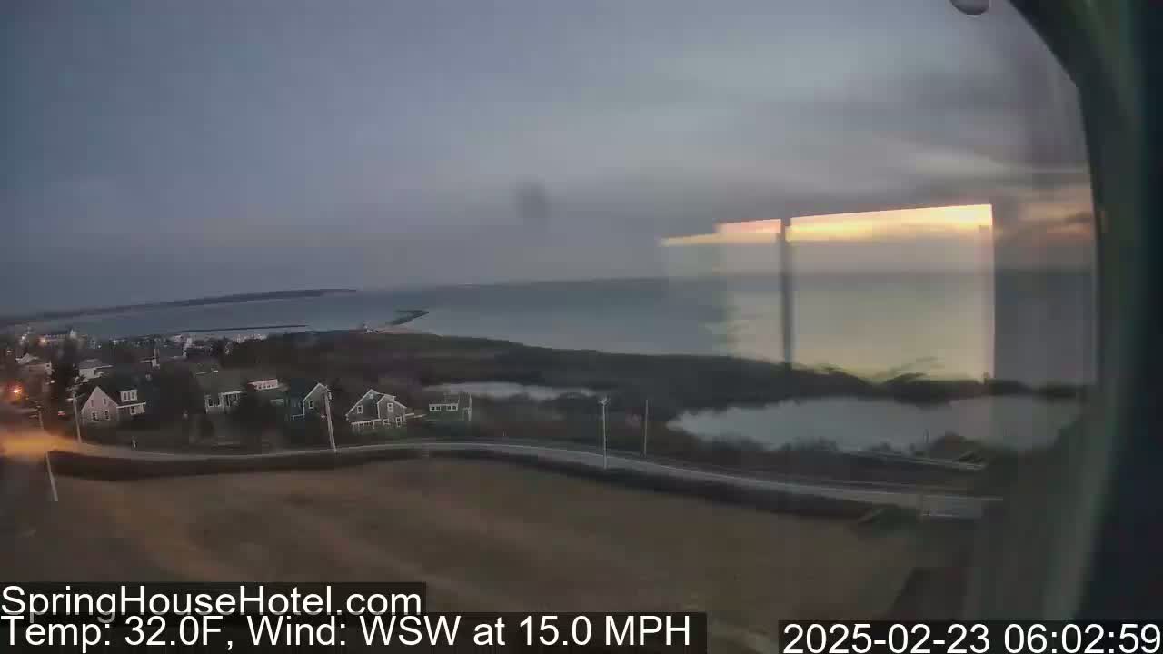 A coastal scene at dawn shows a calm body of water, a residential area with houses, and a road with a 15-mph wind from the west-southwest, and a temperature of 32 degrees Fahrenheit.