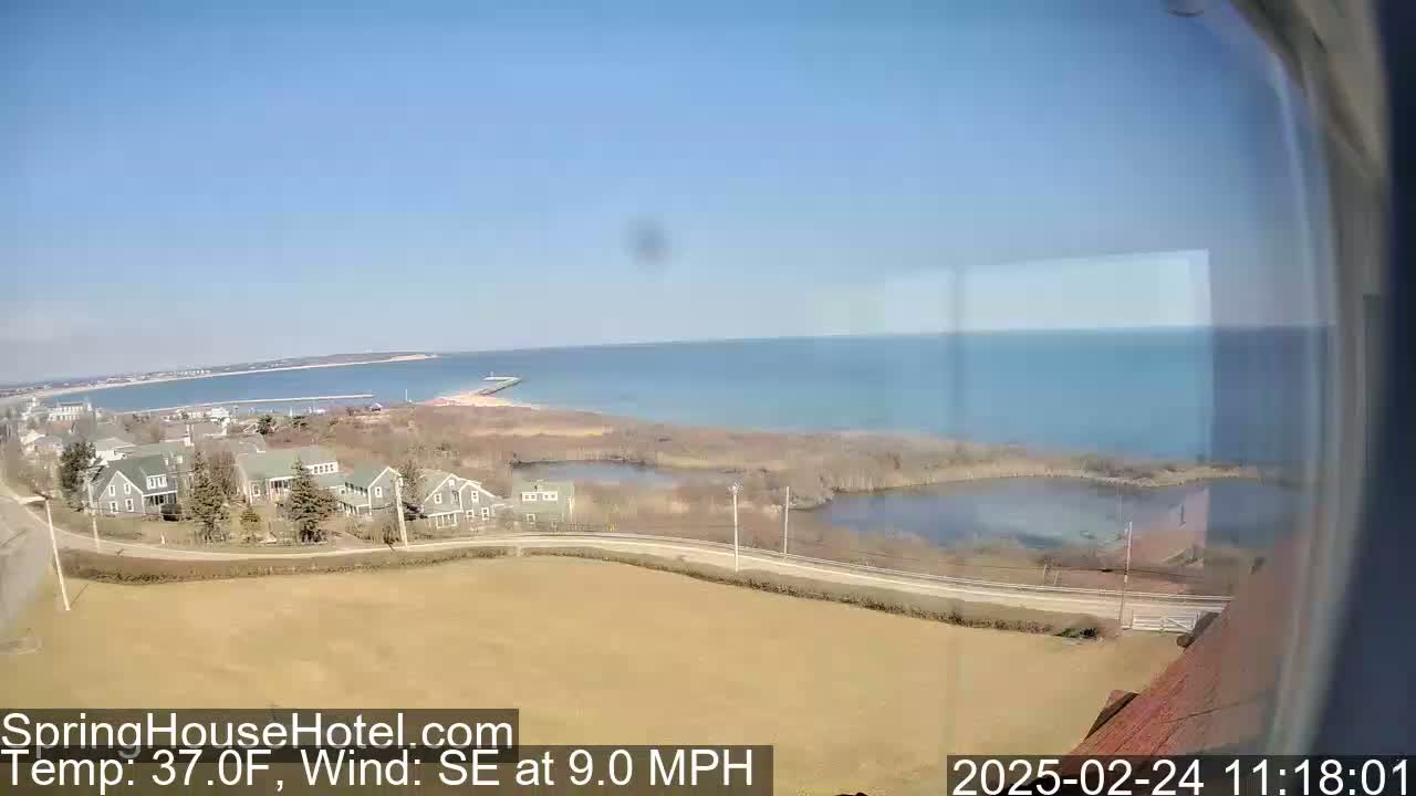 A coastal scene on a clear, sunny day shows a calm body of water, a shoreline with houses and marshland, and a distant landmass under a pale blue sky.