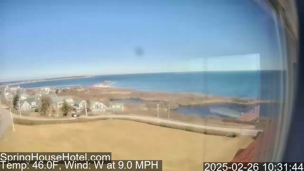 A coastal scene on a clear, sunny day with a 9 mph westerly wind shows a calm body of water, houses, and marshland.