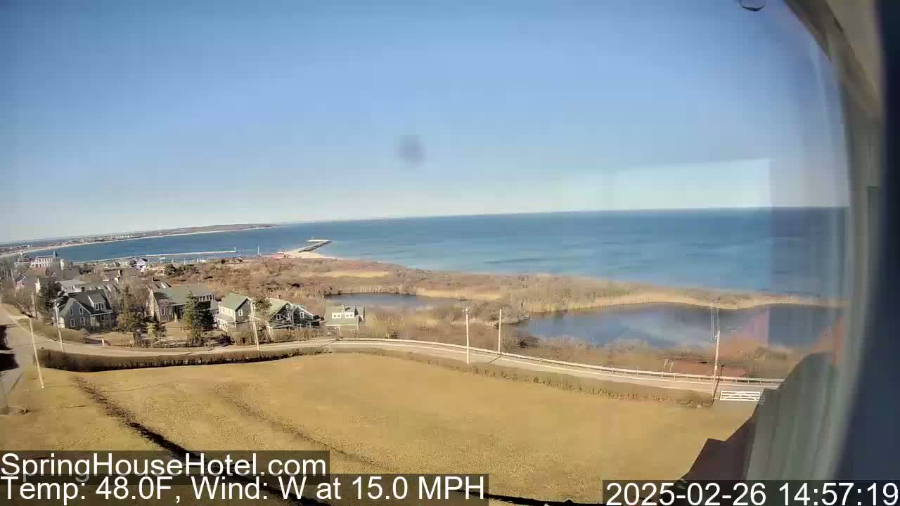 A high-angle view shows a coastal scene on a clear, sunny day with a 15 mph westerly wind, featuring houses, a road, marshland, a calm ocean, and a distant peninsula.