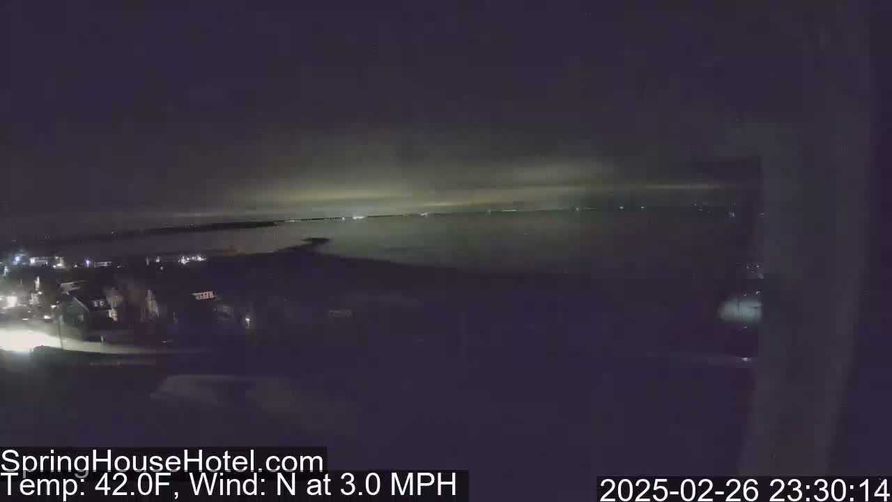 A nighttime, elevated view shows a dark body of water under a partly cloudy sky, with dimly lit houses and a road visible in the foreground.