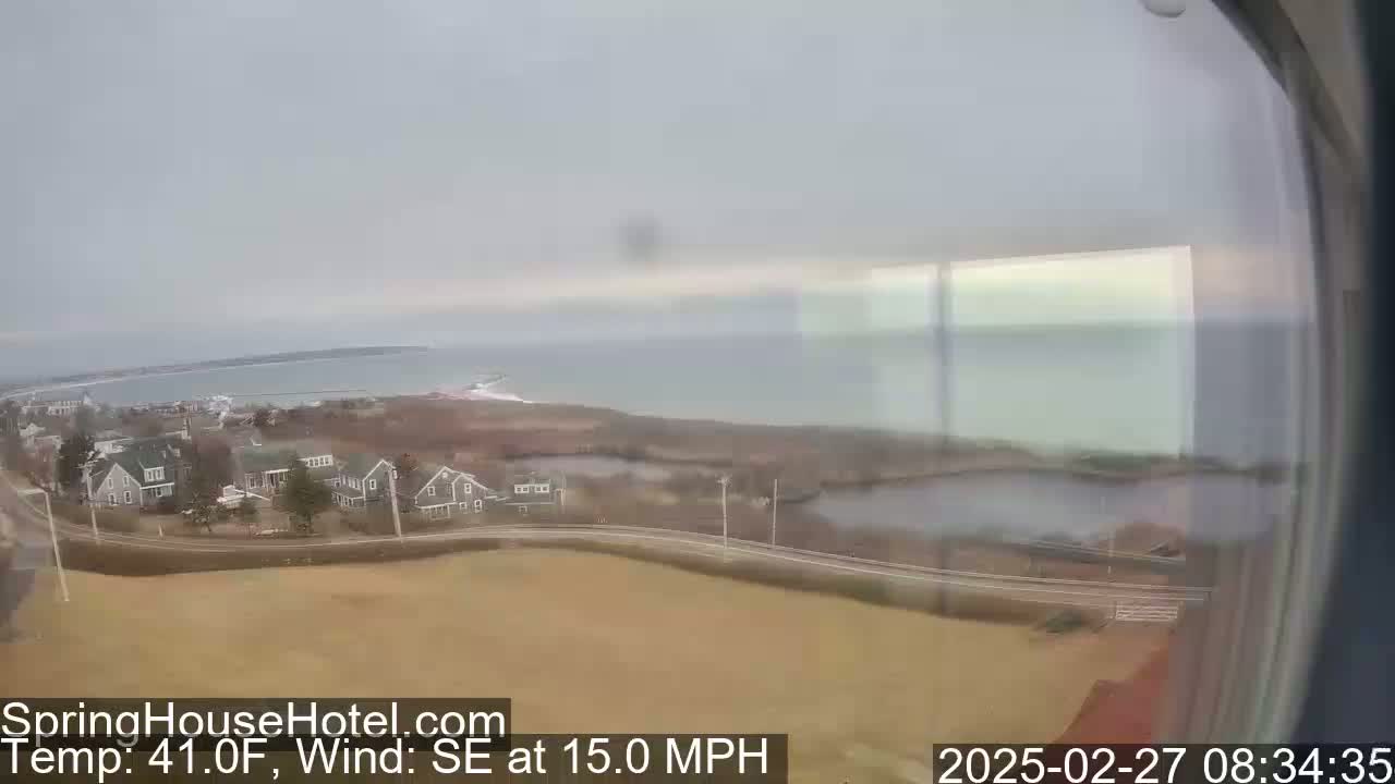 A coastal town with houses is visible under an overcast sky, with a calm body of water in the background and a 15 mph southeast wind.
