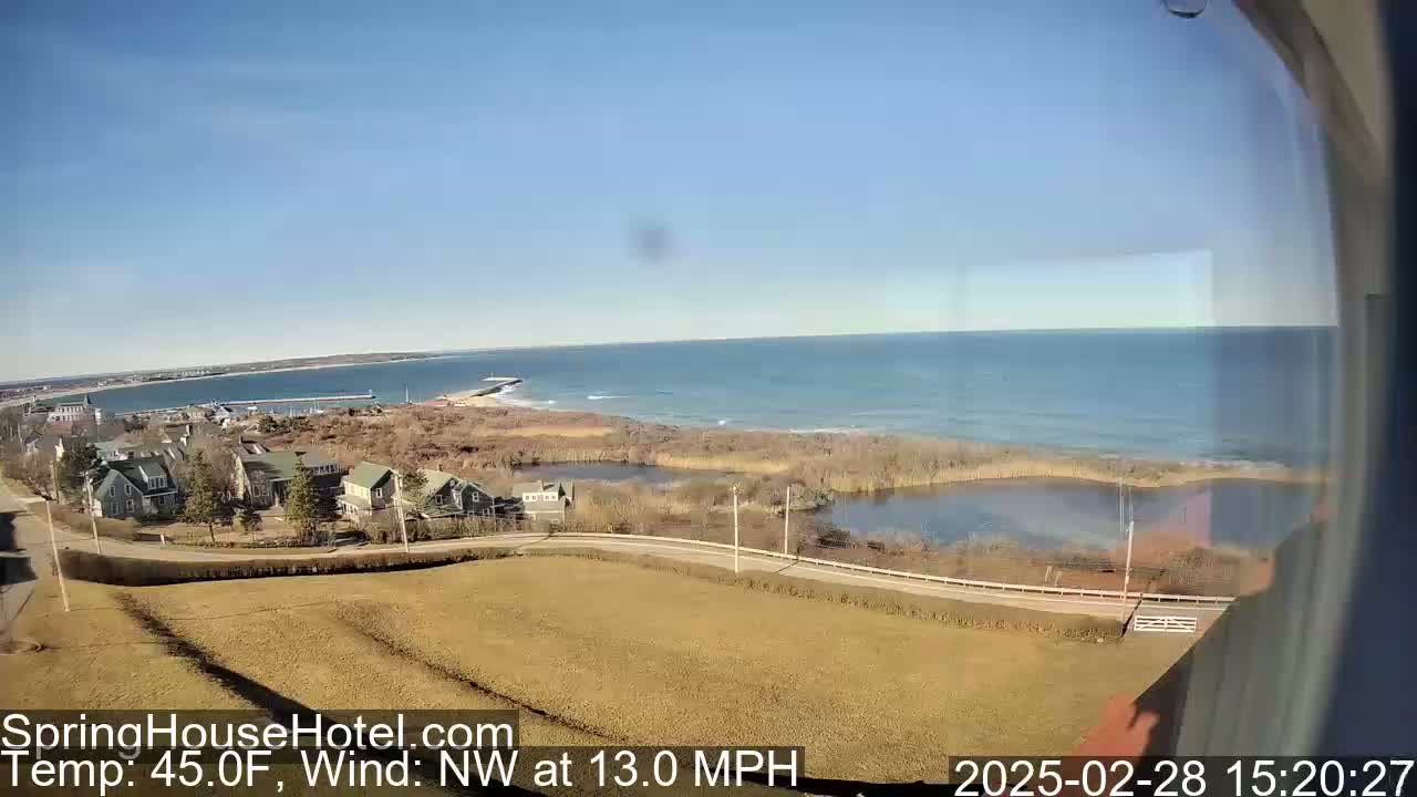 A coastal scene on a clear, sunny day with a northwest wind of 13 mph shows a calm ocean, a small pond, residential buildings, and brown fields.