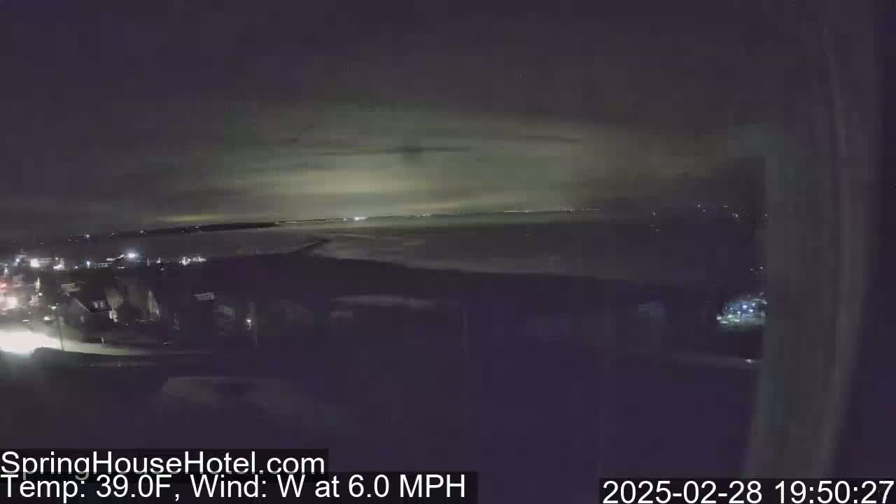 A nighttime view of a town and body of water under a cloudy sky with a 39°F temperature and a 6 mph west wind.