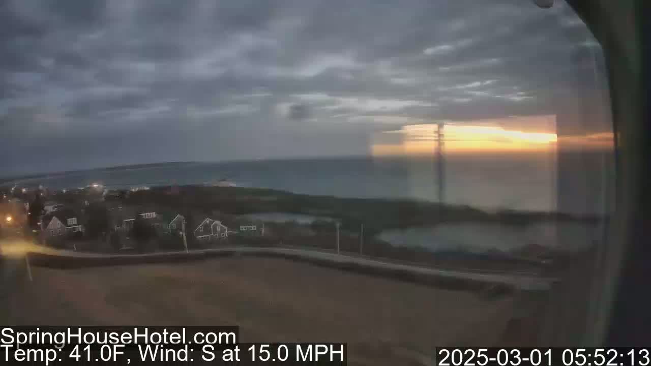 A coastal sunrise with overcast skies and a 15 mph south wind is visible from an elevated vantage point overlooking a residential area and calm ocean waters.