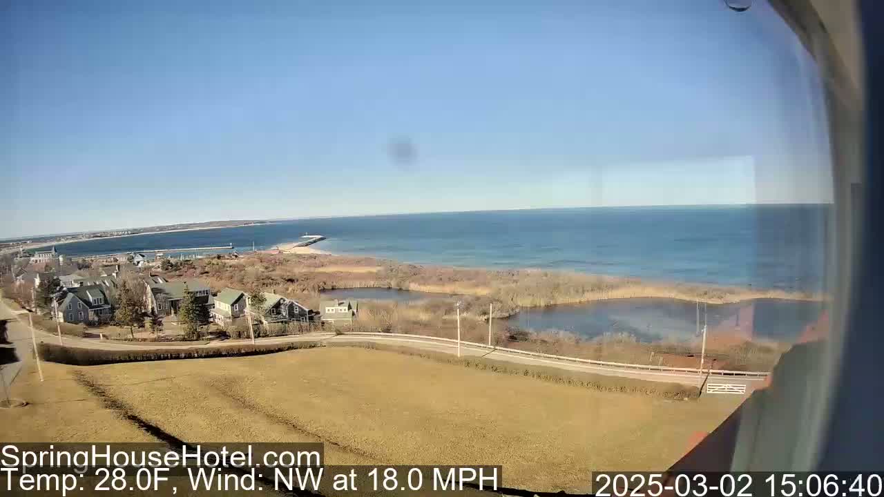 A coastal scene on a clear, sunny day with an 18 mph northwest wind shows houses near a calm body of water that leads to a larger bay or ocean.