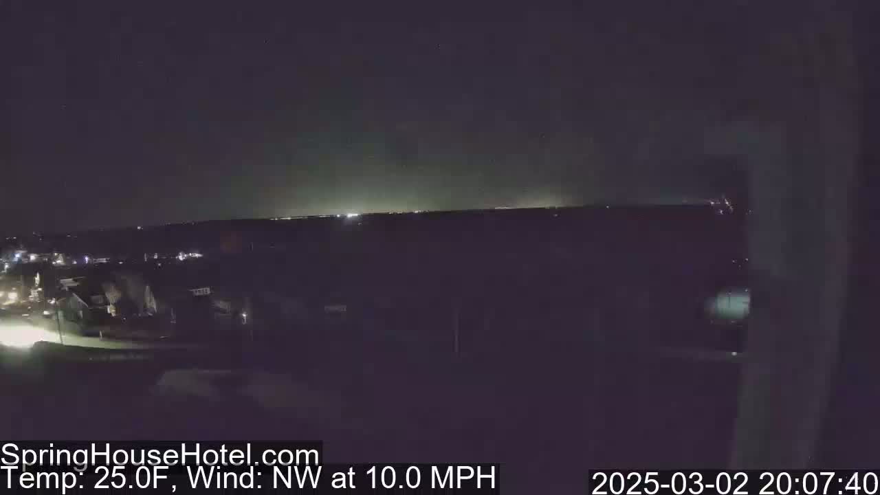 A nighttime view of a suburban area with a distant city skyline illuminated, under a clear sky with a northwest wind at 10 mph and a temperature of 25 degrees Fahrenheit.