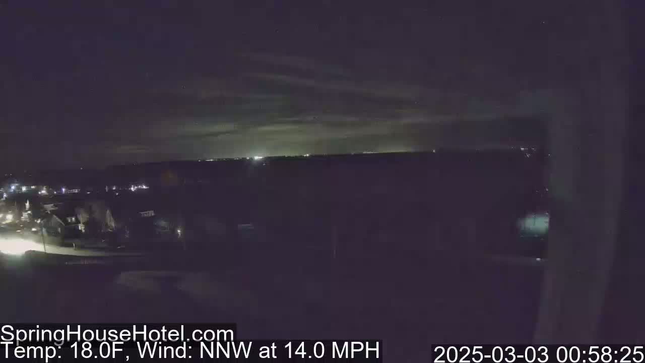 A nighttime view of a residential area with a distant city skyline under a partly cloudy sky, with a 18°F temperature and a 14 mph north-northwest wind.