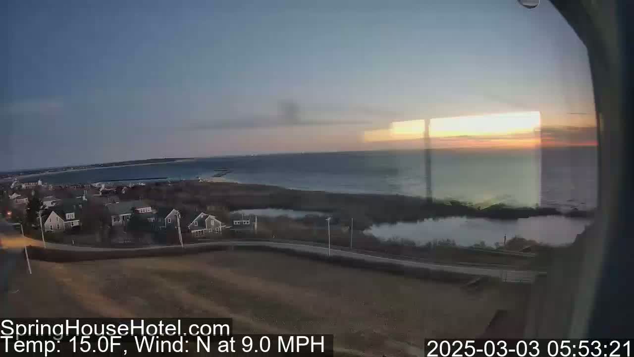 A coastal sunrise over a calm ocean, viewed from above a small town with houses and a road, with a northerly wind at 9.0 mph and a temperature of 15 degrees Fahrenheit.