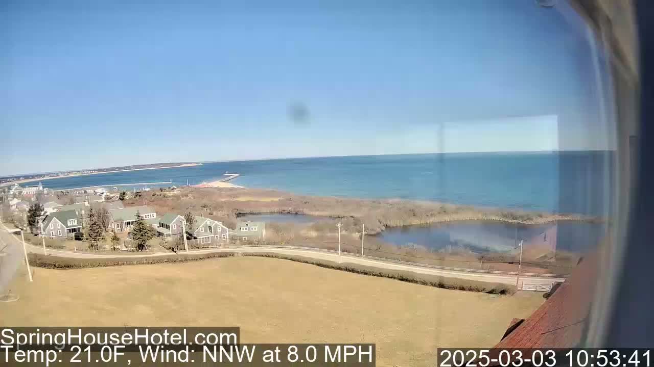 A coastal view shows a calm ocean under a clear blue sky, with houses and marshland in the foreground, and a wind of 8.0 mph from the north-northwest at a temperature of 21.0°F.