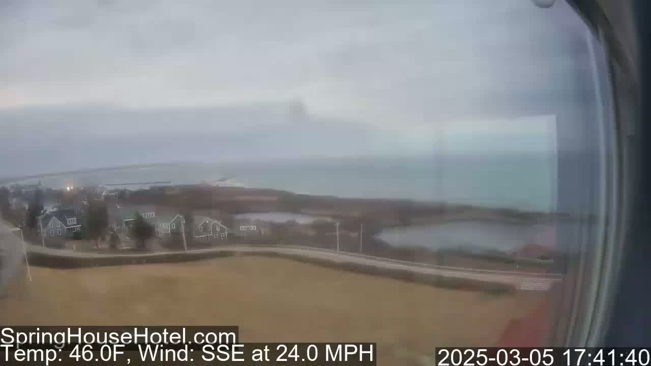 A coastal scene on an overcast day shows a residential area with houses and a body of water, a calm ocean in the distance, and a 24 mph wind from the SSE.