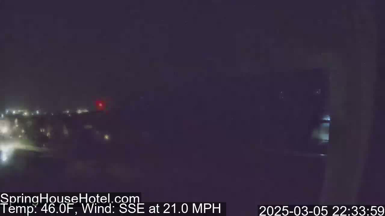 A nighttime view of dimly lit buildings and a dark landscape under a cloudy sky with a 21 mph wind from the SSE at 46°F.