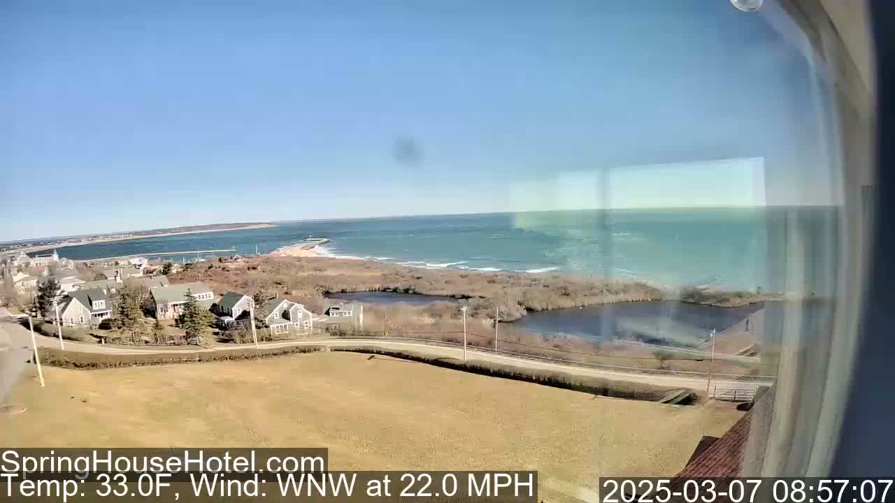 A coastal scene with a calm bay, a windy ocean, and a collection of houses viewed from an elevated position on a clear, sunny day with a 22 mph wind.