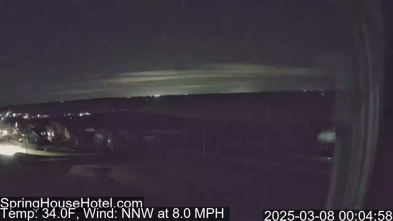 A nighttime long shot of a town under a mostly clear sky with a faint horizon glow, a wind of 8 mph from the north-northwest, and a temperature of 34°F.