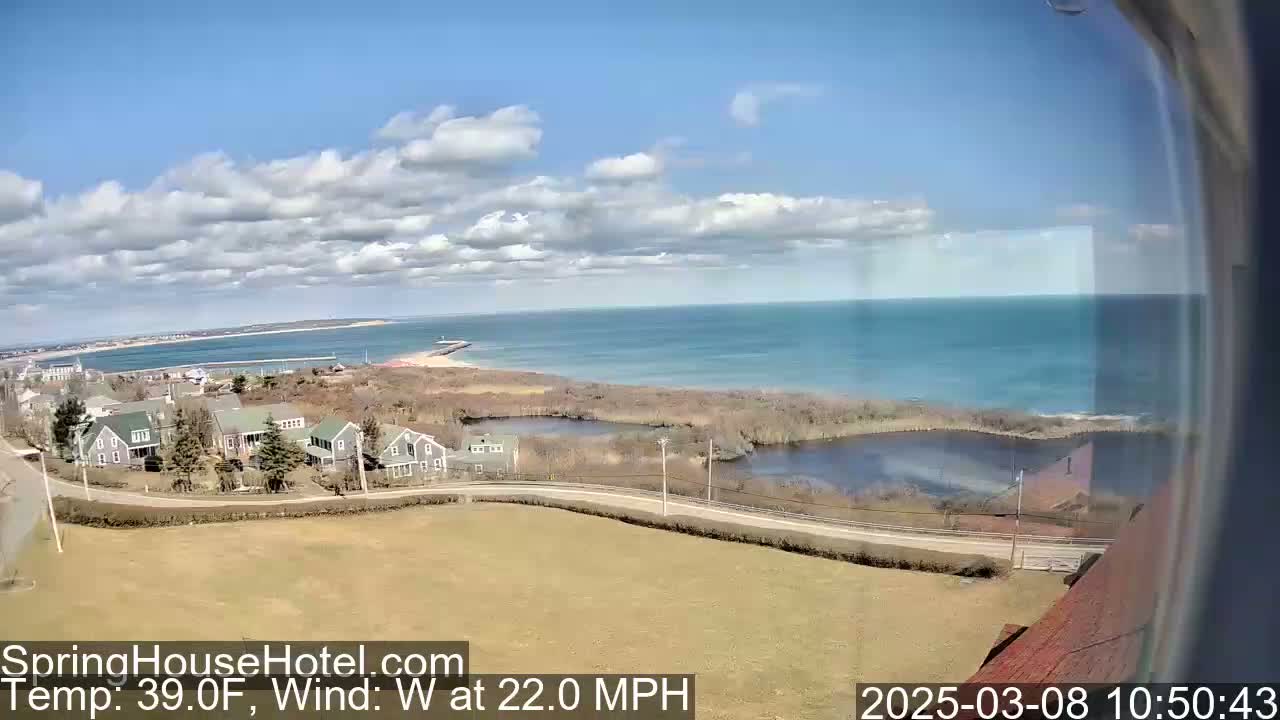 A coastal scene on a partly cloudy day with a west wind at 22 mph shows a tranquil body of water next to a road winding through houses and marshland, with a larger body of water and a distant shoreline visible in the background.