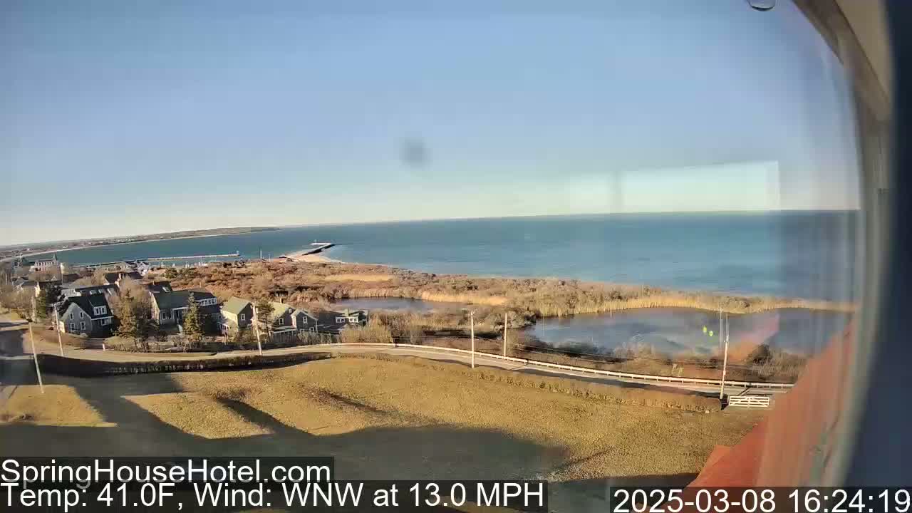 A coastal scene on a sunny day with a 13 mph wind shows houses, a body of water, and a shoreline with a pier.