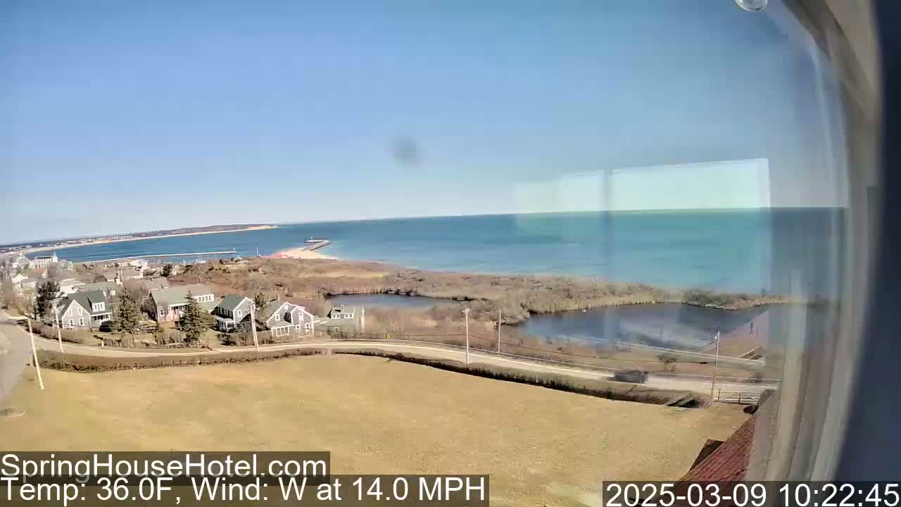 A coastal scene on a sunny day with a 14 mph westerly wind shows a calm body of water, a small town nestled near the shore, and a marshy area separating the town from the water.