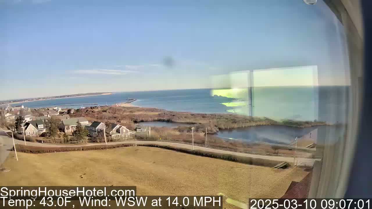 A coastal view shows a town with houses and a calm ocean under a clear blue sky, with a 14 mph wind from the west-southwest.